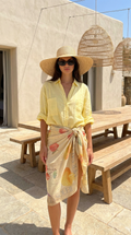 Woman in a yellow shirt and floral skirt standing outdoors with wooden benches and hanging baskets.